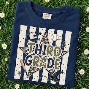 以下が含まれることがあります： 大きな文字で「THIRD GRADE」と書かれたグラフィックプリントが施されたネイビーブルーのTシャツ。中央の星、野球帽、野球ボール、バット、フットボールヘルメットが、縦縞の背景の上にデザインされています。
