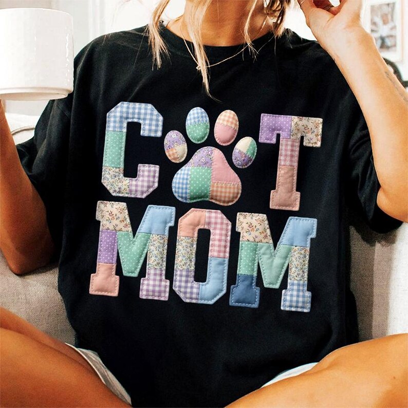 Pode incluir: T-shirt preto com as palavras "CAT MOM" em patchwork e uma pegada. As letras e a pegada s&atilde;o feitas de v&aacute;rios tecidos estampados em cores pastel.