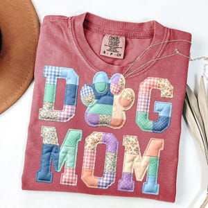 以下が含まれることがあります： 「DOG MOM」の文字と足跡がパッチワークで施された赤いTシャツ。文字と足跡は、ギンガムチェックや花柄など、さまざまな柄の生地で作られています。シルバーのネックレスがシャツの上にかけられています。