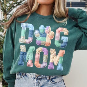 以下が含まれることがあります： 「DOG MOM」の文字と足跡がパッチワークでデザインされた、ティールグリーンのスウェットシャツ。文字と足跡は、青、ピンク、紫の色合いの様々な柄の生地で作られています。スウェットシャツはカジュアルなスタイルです。