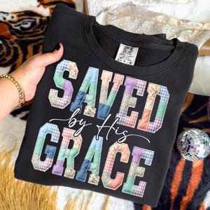 May include: Black sweatshirt with the words "SAVED by His GRACE" in a patchwork design. The letters are made of various pastel-colored fabrics. The sweatshirt has a crew neck and a tag that says "Comfort Colors".