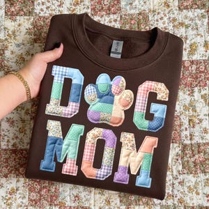 以下が含まれることがあります： 「DOG MOM」の文字と足跡がパッチワークで施された茶色のスウェットシャツ。文字と足跡は、青、緑、ピンク、紫の色合いの様々な柄の生地で作られています。