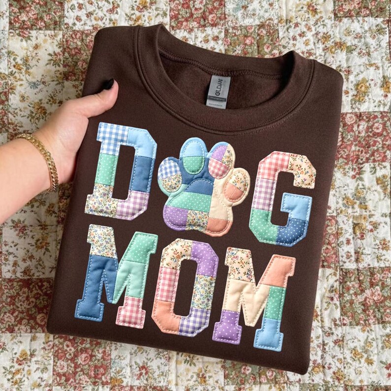 以下が含まれることがあります： 「DOG MOM」の文字と足跡がパッチワークで施された茶色のスウェットシャツ。文字と足跡は、青、緑、ピンク、紫の色合いの様々な柄の生地で作られています。