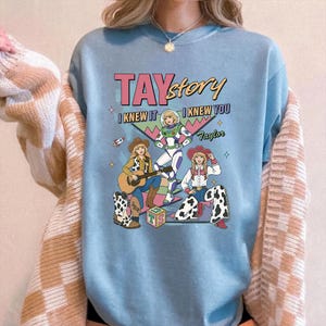 May include: A light blue crewneck sweatshirt featuring a graphic print with the text TAYstory, I KNEW IT, I KNEW YOU, and Taylor. The design includes three illustrated figures in cowboy and space-themed outfits, a guitar, and a block with TS.