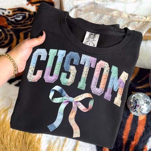 May include: Black sweatshirt with the word "CUSTOM" appliqued in colorful patchwork fabric. A matching patchwork bow is sewn below the text. The sweatshirt has a crew neck and a tag that says "COMFORT COLORS".