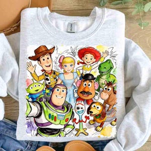 Pode incluir: Camisola cinza claro com um gr&aacute;fico colorido de personagens do Toy Story. O design inclui Woody, Buzz Lightyear, Bo Peep, Jessie, Rex, Sr. Batata e outros. A camisola tem gola redonda.