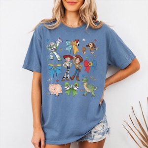 Op de afbeelding: Een blauw t-shirt met korte mouwen en een grafische print van diverse geanimeerde personages, waaronder een space ranger, een cowgirl, een cowboy, een varken, een dinosaurus en een hond, afgewisseld met kleurrijke strikken en kleine sterren.