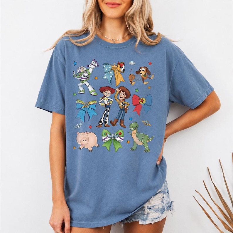 Op de afbeelding: Een blauw t-shirt met korte mouwen en een grafische print van diverse geanimeerde personages, waaronder een space ranger, een cowgirl, een cowboy, een varken, een dinosaurus en een hond, afgewisseld met kleurrijke strikken en kleine sterren.