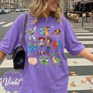Op de afbeelding: Een violetkleurig T-shirt met korte mouwen en een grafische print van diverse tekenfilmfiguren, waaronder een space ranger, een cowgirl, een cowboy, een varken, een dinosaurus en een hond, afgewisseld met kleurrijke strikken en sterren.