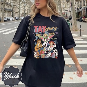 May include: A black t-shirt featuring a graphic print with the text TAY story I KNEW IT I KNEW YOU Taylor. The design includes three illustrated figures in cowboy and space-themed outfits, a guitar, a toy block, and colorful geometric shapes.