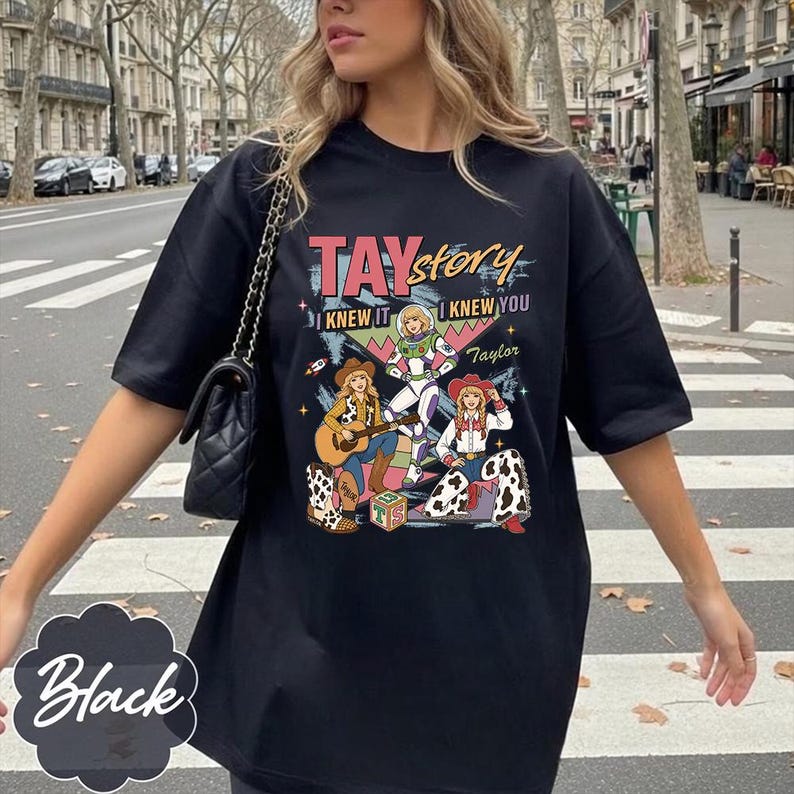 May include: A black t-shirt featuring a graphic print with the text TAY story I KNEW IT I KNEW YOU Taylor. The design includes three illustrated figures in cowboy and space-themed outfits, a guitar, a toy block, and colorful geometric shapes.