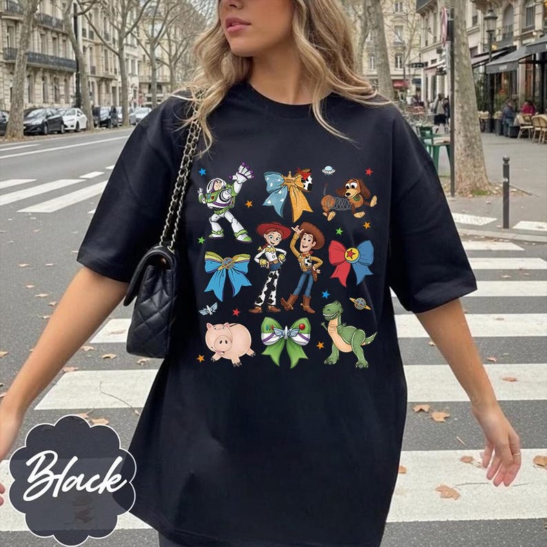 Op de afbeelding: Een zwart t-shirt met korte mouwen en een grafische print van diverse Toy Story-personages, waaronder Buzz Lightyear, Woody, Jessie, Slinky Dog, Rex en Hamm, afgewisseld met kleurrijke strikken en kleine sterretjes op de borst.