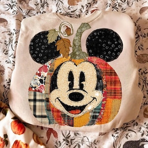 May include: Beige sweatshirt featuring a Mickey Mouse pumpkin design. The pumpkin is made of patchwork fabric in various colors and patterns, with black ears and a green stem. A festive fall-themed apparel item.