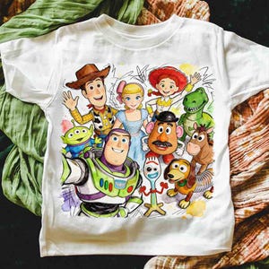 Pode incluir: Camiseta branca com uma ilustra&ccedil;&atilde;o colorida em aquarela dos personagens de Toy Story. O design inclui Woody, Buzz Lightyear, Bo Peep, Jessie, Rex, Sr. Batata, Slinky Dog e outros personagens do filme.