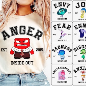 May include: A white t-shirt with a black and white graphic of the character Anger from the movie Inside Out. The graphic is in the style of a college varsity logo with the text "Anger" in a black arch above the character and "EST 2015" below the character. The text "Inside Out" is below the character.