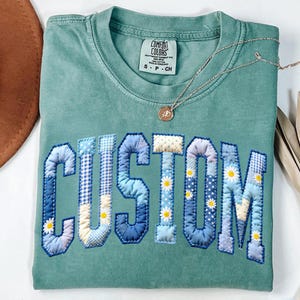 May include: A seafoam green t-shirt with the word "CUSTOM" appliqued in a patchwork design. The letters feature various blue patterns and small daisy accents. A silver necklace with a pendant rests on the shirt.