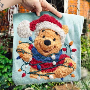 Puede incluir: Cojín cuadrado bordado con Winnie the Pooh con gorro de Papá Noel y luces navideñas. El oso es marrón con una nariz negra y lleva un jersey rojo, azul y blanco. El fondo es azul claro.