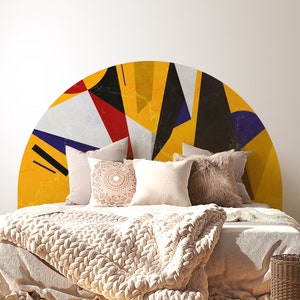 May include: A bed with a white headboard and a yellow, blue, red, and black geometric pattern headboard decal. The bed has a white duvet and a beige knitted throw blanket. A brown wicker basket is on the floor next to the bed. A white lamp with a beige lampshade is on a wooden side table.