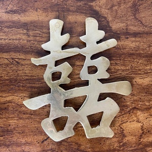 May include: A brass Chinese character for "happiness" or "good fortune". The character is made of a single piece of metal and has a slightly aged finish.