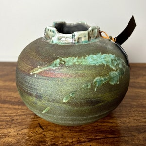 May include: A small, round ceramic vase with a green and brown glaze. The vase has a unique, textured rim with a raised design.