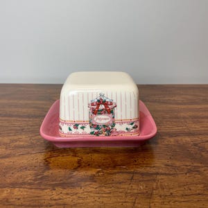 May include: A white ceramic butter dish with a pink floral design and a pink lid. The lid has a pink and white striped pattern and a floral design with the word "Purpose" on it.