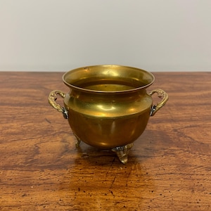 May include: A small, round brass pot with three feet and two ornate handles. The pot has a wide opening and a polished, reflective surface. The brass has a warm, golden hue, and the pot appears to be an antique or vintage item.