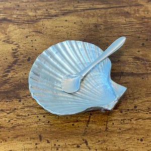May include: A silver-coloured seashell-shaped dish with a small spoon inside. The dish has a textured, ridged surface, and the spoon is smooth. The items are likely for serving condiments or small portions.