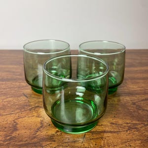 May include: Three green glass tumblers with a rounded bottom and a slightly flared lip. The glasses are sitting on a wooden surface.