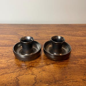 May include: Two black metal candle holders with a smooth, slightly textured finish. The holders are round and have a raised lip around the top.