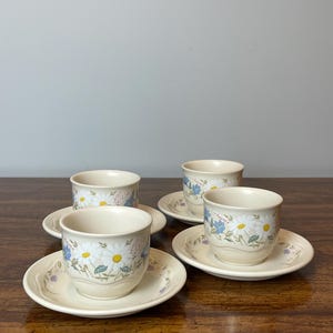May include: Four cream-colored teacups and saucers with a daisy floral pattern. The cups and saucers are arranged on a dark wooden surface. The daisy design features white petals, yellow centers, and blue accents.