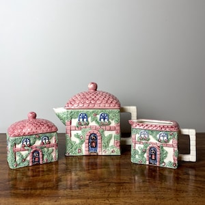 Könnte beinhalten: Ein rosa-grünes Keramik-Set aus Teekanne, Zuckerdose und Milchkännchen in Form von Häusern mit Efeu und Blumen.