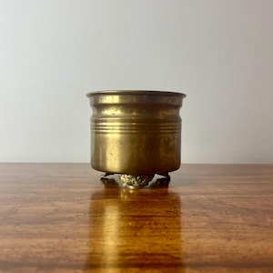 May include: A small, antique brass planter with a cylindrical body and three ornate feet. The planter has horizontal ridges around its midsection and a slightly flared rim. The brass has a warm, aged patina, suggesting its vintage. The planter is empty.