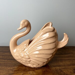 May include: A pale pink ceramic swan planter with a detailed design. The swan's wings are spread out and the head is turned to the side. The planter is perfect for adding a touch of elegance to any home.