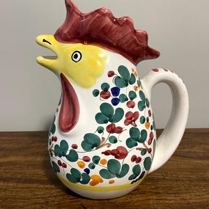May include: A ceramic rooster pitcher with a white base, yellow beak, and a red comb and wattle. The pitcher is decorated with green leaves, red flowers, and colorful berries. The handle is white with red polka dots.