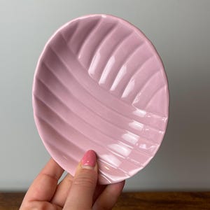 May include: A small, round, pink ceramic dish with a ridged, leaf-like design. The dish is held in a hand, showcasing its delicate form and colour. The dish is a soft, pastel pink.