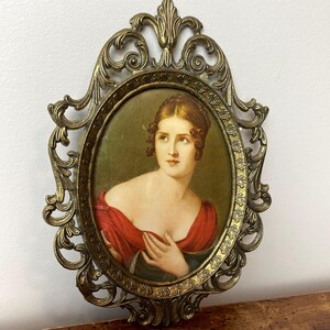 May include: A vintage oval portrait of a woman in a red dress, framed in a gold ornate metal frame.