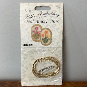 May include: A Bucilla Silk Ribbon Embroidery kit for Oval Brooch Pins. The package contains a gold-coloured oval brooch and a card with two embroidered oval designs of tulips in pink, orange, and yellow.