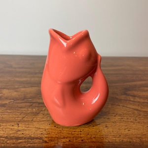May include: A coral pink ceramic vase shaped like a fish. The vase has a small opening at the top and a handle on the side.