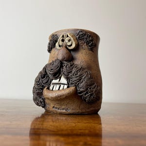 May include: A brown ceramic mug with a face sculpted on the front. The face has a large mustache, curly hair, a nose, and teeth. The mug is sitting on a wooden surface.