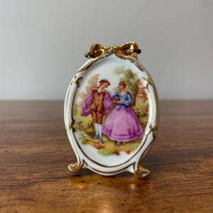 May include: A small porcelain picture frame with a gold trim and a painted scene of a couple in a garden. The frame has a bow at the top and three feet.