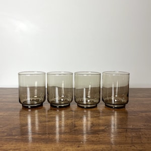 May include: Set of four clear glass tumblers with a brown tint. The glasses are arranged in a row on a wooden surface.
