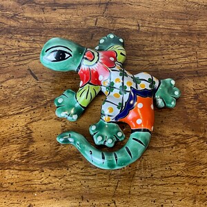 May include: A colorful ceramic lizard figurine with a green head and tail, and a body decorated with red, white, orange, and blue patterns. The lizard has detailed feet and a whimsical design, perfect for home decor.