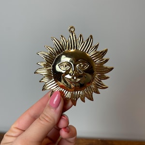 May include: A gold-colored sun ornament with a smiling face. The sun has a raised, textured design and is made of metal.