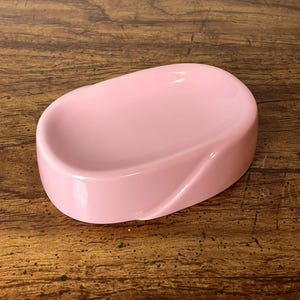 May include: A pink ceramic soap dish with a smooth, oval shape. The dish has a slight curve on the bottom edge.