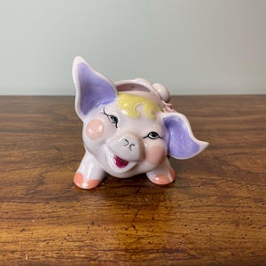 May include: A ceramic pig ornament in pastel pink with lavender ears and yellow accents. The pig has rosy cheeks, a smiling mouth, and black eyelashes. The figurine is a decorative item.