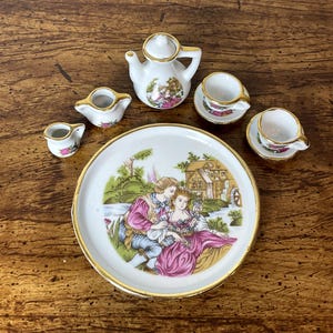Puede incluir: Un juego de té de porcelana en miniatura con ribete dorado. El juego incluye una tetera, una jarra de crema, un azucarero, dos tazas de té y dos platillos. La tetera presenta una escena pintada de una pareja en un jardín. Las tazas de té y los platillos tienen un diseño floral.
