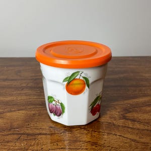 Peut inclure: Un pot en céramique blanche avec un couvercle orange vif. Le pot a un motif de fruits avec une orange, des prunes et des cerises.