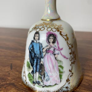 May include: A white porcelain bell with a gold trim and a painted scene of a couple in 18th-century clothing. The bell has an iridescent glaze.