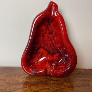 May include: A red ceramic pear-shaped dish with a glossy finish. The dish has a deep, rounded interior and a smooth, slightly textured exterior.