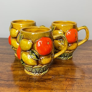 Op de afbeelding: Drie keramische mokken met een citrusfruitontwerp. De mokken zijn geel met oranje en groene accenten. De mokken staan op een houten oppervlak.